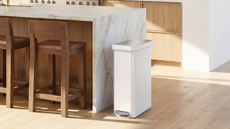 A sleek white pedal trash can beside a marble kitchen island in a modern luxury kitchen, featuring warm wood cabinetry, dark walnut bar stools, light oak flooring, and soft natural sunlight.