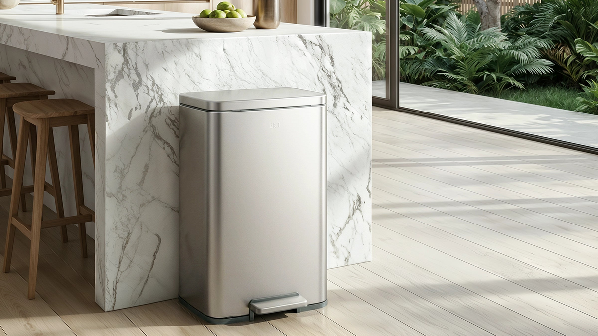 A sleek stainless steel pedal trash can beside a white marble kitchen island in a modern luxury home, featuring light oak flooring, wooden bar stools, floor-to-ceiling glass doors, and lush tropical greenery in natural sunlight.