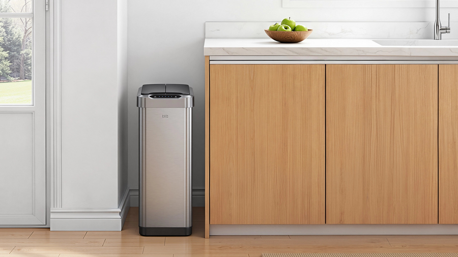 A sleek stainless steel EKO motion-sensor trash can beside light oak kitchen cabinetry, featuring a white quartz countertop, a bowl of green apples, warm wood flooring, and a bright, minimalist modern kitchen with natural daylight.