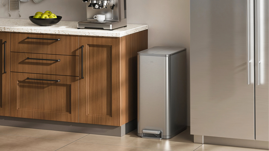 A sleek stainless steel step trash can beside a modern refrigerator in an elegant kitchen, featuring rich walnut cabinetry, a light stone countertop, a coffee machine, and warm natural lighting.