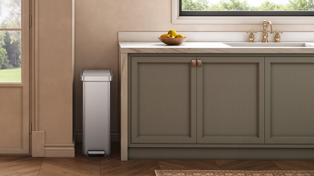 Stainless steel step trash can beside kitchen cabinets in a modern kitchen with warm neutral tones and wood flooring.