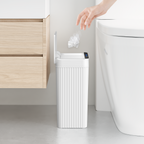 White trash can next to a toilet with a hand disposing of trash, in a bathroom setting.