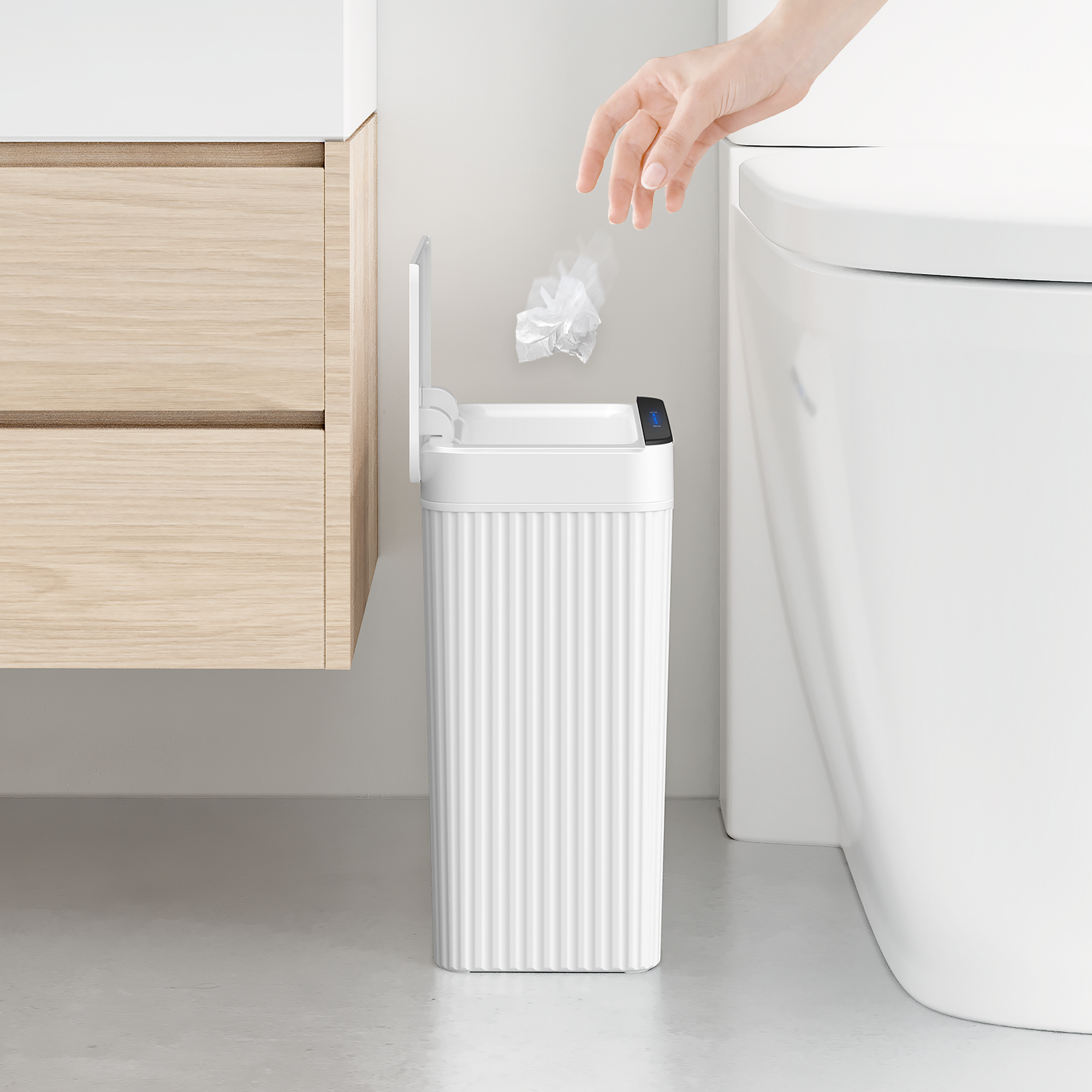 White trash can next to a toilet with a hand disposing of trash, in a bathroom setting.