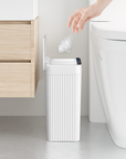 White trash can next to a toilet with a hand disposing of trash, in a bathroom setting.
