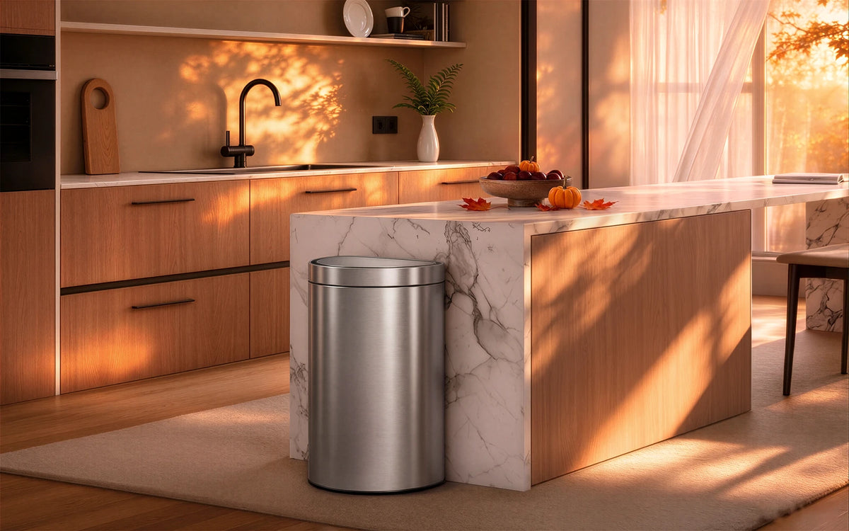 A stainless steel semi-round sensor trash can stands in a warm, sunlit kitchen next to a white island. The scene is decorated for autumn, with candles, pumpkins, and warm light streaming through French doors in the background.